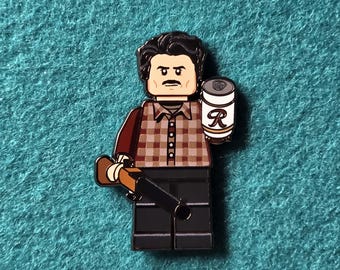Op de afbeelding: Een Lego-stijl pin met een personage in een bruin en beige geruit overhemd, zwarte broek en een donkere snor. Het personage houdt een blikje vast met een wit label en een grote "R", en een bruin voorwerp dat op een geweer lijkt.