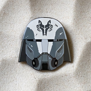 May include: A gray and white helmet-shaped pin with a wolf emblem on top. The pin has a detailed design with various shades of gray and black, and is set against a sandy background.