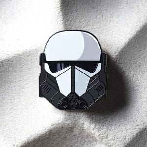 Puede incluir: Un pin de esmalte negro, blanco y gris con un diseño de casco estilizado. El pin tiene un acabado brillante y está sobre un fondo neutro. El diseño del casco tiene una forma angular distintiva.