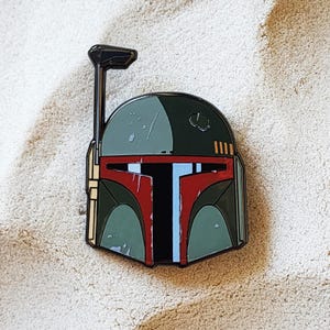 May include: Enamel pin featuring the helmet of a bounty hunter. The pin has a black outline with a green, red, and blue color scheme. The helmet has a black antenna and is set against a sandy background.