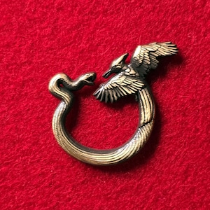 May include: A bronze-toned ring featuring a snake and a bird of prey facing each other. The snake curves around the ring, while the bird has outstretched wings. The ring is set against a red background.