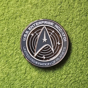 Könnte beinhalten: Ein runder, silberfarbener Anstecker mit dem Text "U.S.S. ENTERPRISE NCC-1701" und "UNITED FEDERATION OF PLANETS", der ein graues und weißes Starfleet-Abzeichen umrahmt. Der Anstecker befindet sich auf einem grünen Hintergrund.