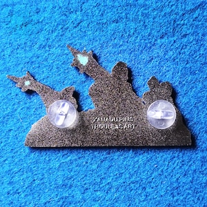 Pode incluir: Um alfinete de esmalte cinza escuro com um design de silhueta de duas figuras apontando para cima. O alfinete tem duas embreagens de borracha transparentes na parte de tr&aacute;s. O texto "XAIADUPINS THOULESSART" est&aacute; impresso no alfinete.