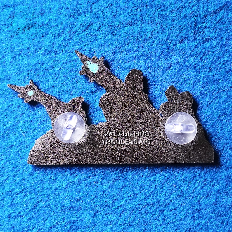 Pode incluir: Um alfinete de esmalte cinza escuro com um design de silhueta de duas figuras apontando para cima. O alfinete tem duas embreagens de borracha transparentes na parte de tr&aacute;s. O texto "XAIADUPINS THOULESSART" est&aacute; impresso no alfinete.