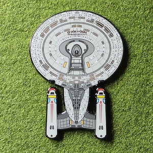 Puede incluir: Un pin detallado de la nave estelar U.S.S. Enterprise NCC-1701-D de Star Trek. El pin presenta un diseño gris y negro con acentos rojos, amarillos y azules. El pin está sobre un fondo verde.