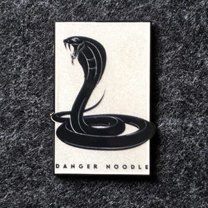 Pode incluir: Gráfico retangular com fundo branco sujo, apresentando uma ilustração de uma cobra negra. A cobra está enrolada e em pé, com o capuz aberto e presas visíveis. As palavras "DANGER NOODLE" estão impressas em preto na parte inferior.