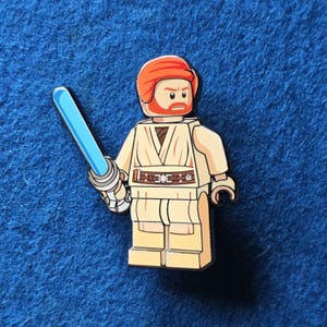 May include: A pin depicting a Lego-style figure with a red helmet, orange beard, and a tan outfit. The character holds a blue lightsaber. The pin is set against a blue background.
