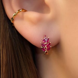 Può includere: Un orecchino a cerchio dorato e un piccolo orecchino a cerchio dorato ornato da un gruppo di pietre preziose rosse rubino taglio marquise. L'orecchino è a forma di piccola foglia o ramo.