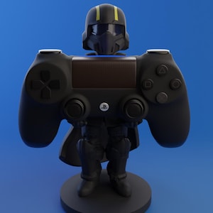 Op de afbeelding: Een zwarte PlayStation 4 controllerstandaard in de vorm van een futuristische soldaat. De standaard heeft een helm, harnas en een cape, waarbij de controller het lichaam vormt. De standaard staat op een ronde zwarte basis, tegen een blauwe achtergrond.
