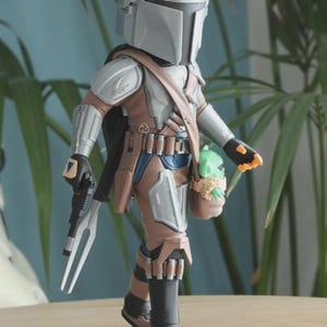 May include: A Mandalorian action figure in a silver helmet and armor, holding a blaster and a small green creature in a brown satchel. The figure is wearing brown and blue clothing and black boots. The figure is standing on a wooden surface.