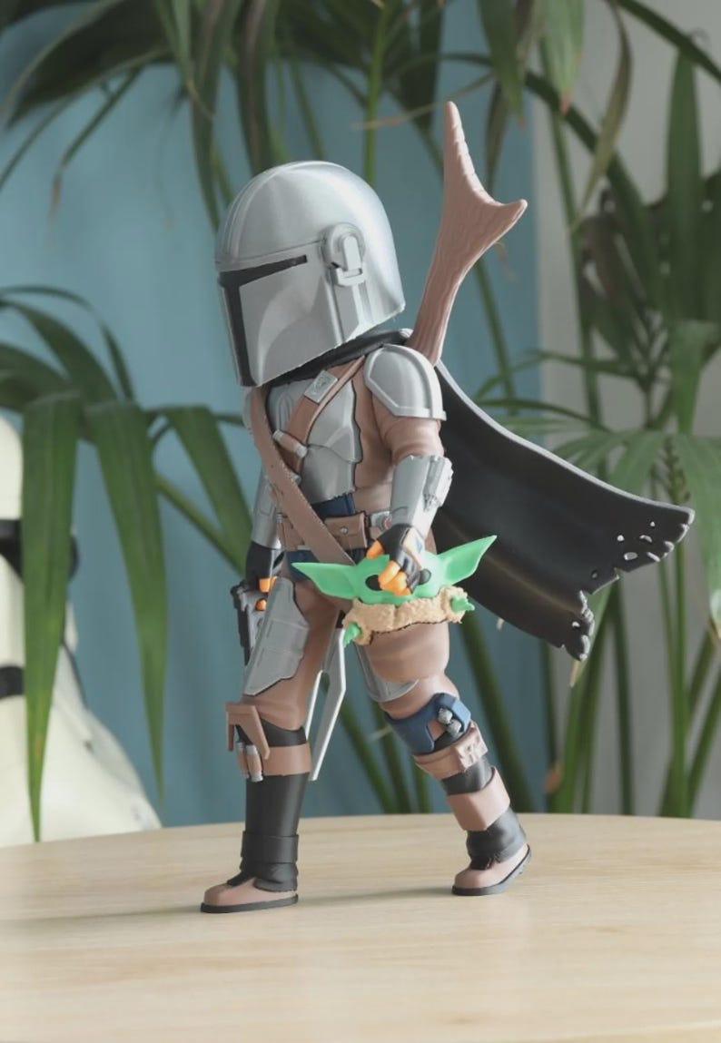 May include: A figurine of a character from a science fiction series, wearing a silver helmet and armor, with a black cape and a green creature. The figure is standing on a wooden surface.