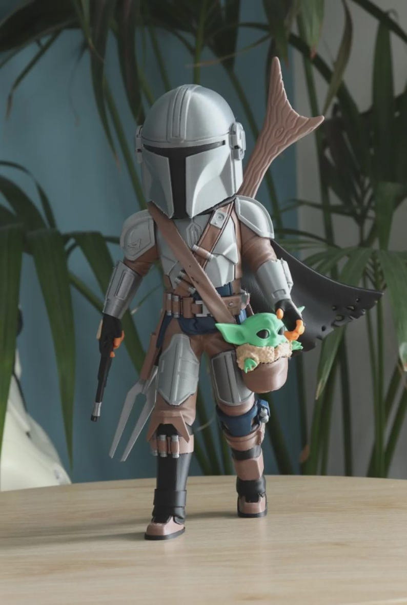 May include: A detailed figurine of The Mandalorian, clad in silver armor, holding a blaster and a fork-like weapon. A small green Grogu figure is nestled in a pouch. The figure has a brown cape and a wooden spear-like attachment on its back.