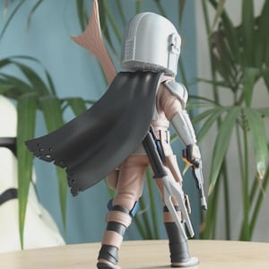 May include: A detailed action figure of a character from a science fiction series. The figure is wearing a silver helmet, a black cape, and tan and gray armor. It is holding two blasters and a spear-like weapon. The figure is standing on a wooden surface.