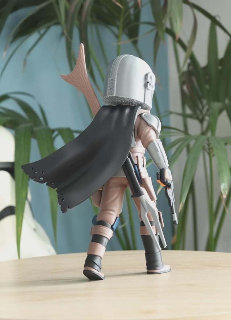 May include: A detailed action figure of a character from a science fiction series. The figure is wearing a silver helmet, a black cape, and tan and gray armor. It is holding two blasters and a spear-like weapon. The figure is standing on a wooden surface.