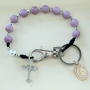 May include: A rosary-style keychain with light purple beads, a silver cross pendant, and a Virgin Mary charm. The keychain has a clasp and a key ring for attaching keys or other items.