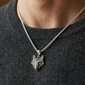 Può includere: Collana color argento con un ciondolo a forma di testa di lupo dettagliata. Il ciondolo presenta elementi di design intricati, sospesi da una delicata catena. La collana è indossata su un maglione grigio scuro.