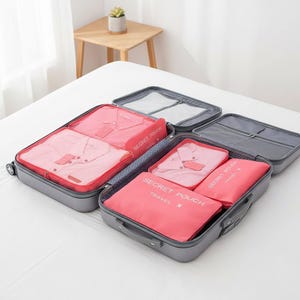Peut inclure: Une valise grise ouverte avec plusieurs compartiments, remplie de cubes d'emballage roses et de chemises pliées. Les cubes d'emballage portent l'inscription "SECRET POUCH TRAVEL". La valise est ouverte sur une surface blanche.
