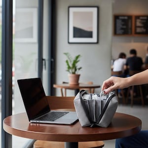 May include: A grey fabric tech organiser pouch with a zippered opening and internal dividers, containing various white charging cables and electronic accessories, positioned next to an open silver laptop on a round wooden table.