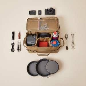 May include: A tan canvas bag containing camping and photography equipment. Includes a camera, cooking pots, a flashlight, a multi-tool, and a small propane stove. Also includes a set of three dark grey cooking pans.