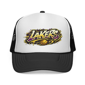 Op de afbeelding: Een zwart-witte truckerpet met het Lakers-logo. Het logo toont het woord "Lakers" in goud met paarse accenten, een basketbal en een druppeleffect. De pet heeft een wit voorpaneel en een zwarte klep en mesh achterkant.