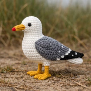 May include: A crocheted seagull toy. The bird has a white body, grey wings, and a black and white tail. It features a yellow beak with a red tip and yellow feet. The seagull is standing on a sandy surface.