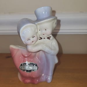 May include: A ceramic figurine of a couple embracing, with a top hat and bonnet. The figurine is painted in pastel colors, with a pink and purple base. The text "Souvenir of LANSING, MICH" is visible.