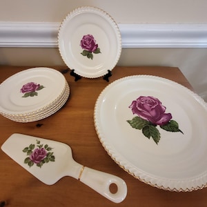 May include: A cream-colored porcelain dish set featuring a purple rose design. The set includes plates, a serving platter, and a cake server. Each piece has a gold trim and a scalloped edge, creating an elegant aesthetic.