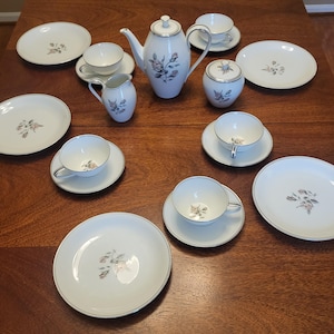 May include: A white porcelain tea set with a floral design and silver trim. The set includes a teapot, sugar bowl, creamer, cups, saucers, and plates. The floral pattern features pink and gray flowers.
