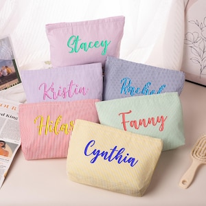 May include: A collection of six rectangular fabric pouches featuring vertical pinstripes in various pastel and bold colors. Each pouch is embroidered with a unique name in a cursive script, including Stacey, Kristin, Rachel, Hilary, Fanny, and Cynthia.