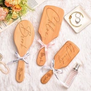 May include: Three light brown wooden hairbrushes with engraved names and floral designs. Each brush is tied with a ribbon. The brushes feature the names Brook, Chloe, and Wendy, along with engraved flower illustrations on the handles.