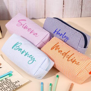 Könnte beinhalten: Vier personalisierte Federmäppchen in den Farben Lavendel, Blau, Marine und Orange. Jedes Etui hat einen Namen in einer Kontrastfarbe gestickt. Die Etuis sind gestreift und haben einen Reißverschluss. Schreibwaren sind um die Etuis herum verstreut.