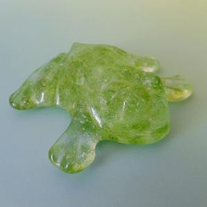 May include: A translucent, lime green glass sculpture with an organic, abstract shape. The piece has a textured surface and a few rounded protrusions, resembling a stylized form. The object is set against a light blue background.