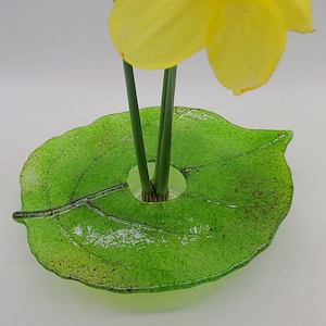May include: A green glass vase shaped like a leaf with a hole in the center for flowers. The vase has a speckled texture and is filled with three yellow daffodils.