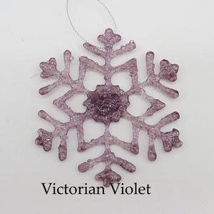 May include: A purple glass snowflake ornament with a glittery finish. The ornament is hanging from a silver string and has the text "Victorian Violet" printed below it.