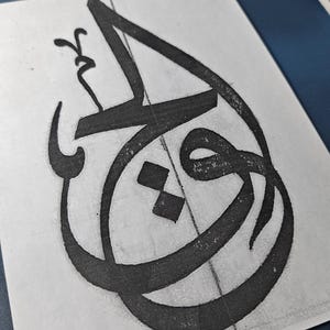 以下が含まれることがあります： 白い紙に黒いアラビア書道のデザインをクローズアップ。このアートワークは、大胆で流れるような線と複雑なディテールを特徴とし、青い背景を背景としています。書道は紙の中央に配置され、アーティストのスキルを披露しています。