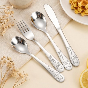 May include: A four-piece stainless steel cutlery set including a knife, a fork, and two spoons. Each piece features the engraved name "Sopia" and a small bear icon on the handle. The utensils have a polished, reflective silver finish.