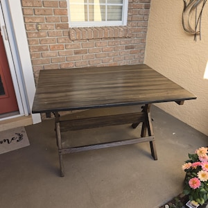 May include: A dark brown, rectangular folding table with a slatted tabletop and trestle-style legs. The table is made of wood and is set up outdoors on a concrete patio. The table measures approximately 30 inches long and 30 inches wide.