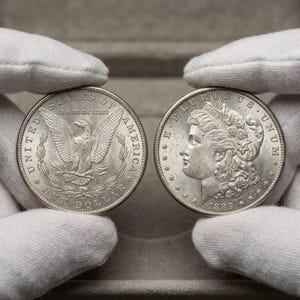 Puede incluir: Dos dólares de plata Morgan, sostenidos por manos enguantadas. Una moneda muestra un águila con las alas extendidas y las palabras "UNITED STATES OF AMERICA" y "ONE DOLLAR". La otra moneda presenta un perfil de mujer y la fecha 1889.
