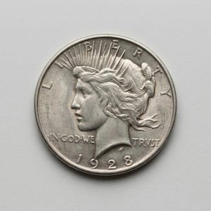 May include: A silver 1923 Peace dollar coin. The obverse features a profile of Lady Liberty with the words "IN GOD WE TRUST" and "LIBERTY". The coin's year, 1923, is visible. The coin is circular and has a detailed design.