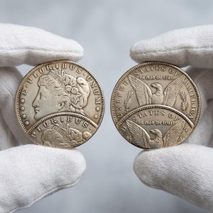 Puede incluir: Dos monedas de tono plateado sostenidas entre dedos con guantes blancos. La moneda de la izquierda muestra un perfil de Lady Liberty con el texto E PLURIBUS UNUM. La moneda de la derecha muestra un águila con las alas extendidas y el texto UNITED STATES OF AMERICA IN GOD WE TRUST.