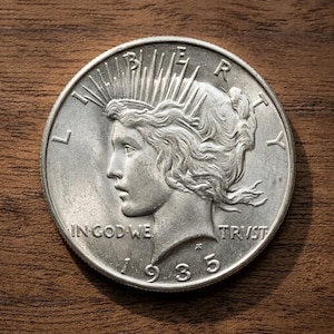 Op de afbeelding: Een zilveren 1935 Peace dollar munt. De voorzijde toont een profiel van Lady Liberty met stralen van haar kroon, en de inscripties "LIBERTY" en "IN GOD WE TRUST". De munt ligt op een houten oppervlak.