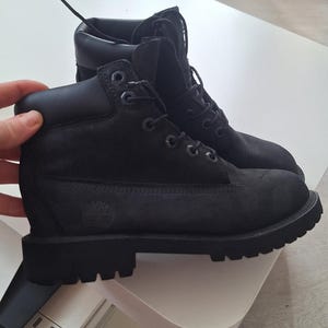 Puede incluir: Un par de botas negras hasta el tobillo con cordones y ojales metálicos, cuello acolchado y una suela de goma gruesa con tacos. El lateral de la bota muestra un logotipo de árbol grabado en relieve, con cordones y costuras de color negro.