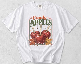 Puede incluir: Una camiseta blanca de manga corta con un estampado gráfico de tres manzanas de caramelo rojas en palitos. El diseño incluye el texto Candy APPLES, una pequeña araña y hojas de otoño, todo dentro de un borde rectangular con detalles naranjas.