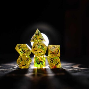 Op de afbeelding: Een verzameling gele harsdobbelstenen met zwarte cijfers, tegen een donkere achtergrond. De dobbelstenen hebben verschillende vormen, waaronder een piramide, een kubus en een 20-zijdige dobbelsteen. Voor tabletop games.