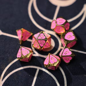 Op de afbeelding: Een set polyhedrale dobbelstenen met een roze hartontwerp en gouden randen. De dobbelstenen zijn genummerd en ontworpen voor tabletop games. Ze liggen op een donkerblauwe ondergrond.