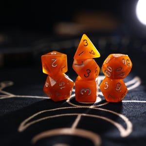 Könnte beinhalten: Ein Set aus sieben halbtransparenten, orangefarbenen polyedrischen Würfeln mit silberfarbenen Zahlen. Die Würfel sind in drei Stapeln angeordnet und umfassen verschiedene Formen, darunter vier-, sechs-, acht-, zehn-, zwölf- und zwanzigseitige Würfel.