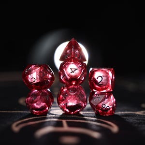 Può includere: Sei dadi poliedrici rossi e traslucidi sono impilati a forma di piramide su uno sfondo scuro. I dadi sono numerati e sembrano essere per giochi da tavolo. I dadi sono illuminati da dietro, creando un effetto luminoso.