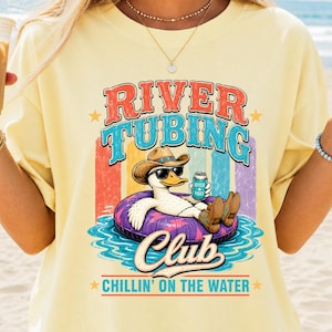 Peut inclure: T-shirt jaune clair avec un motif représentant un canard portant des lunettes de soleil et un chapeau de cowboy, flottant sur un bouée. Le texte indique "River Tubing Club" et "Chillin' on the Water". Le canard tient une canette de "River Vibes".