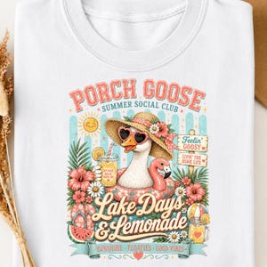 Op de afbeelding: Wit T-shirt met een kleurrijke afbeelding van een gans met zonnebril en strohoed, drijvend op een flamingo-zwemband. Tekst bevat "Porch Goose Summer Social Club" en "Lake Days & Lemonade".