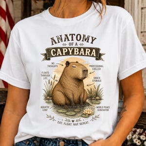 May include: A white t-shirt featuring a brown capybara illustration with the text Anatomy of a Capybara. Labels point to the animal with phrases including No Thoughts, Professional Chiller, Snack Storage, Floats Through Life, Aquatic Loaf, and World Peace Generator, with the text Eat. Float. Nap. Repeat. at the bottom.